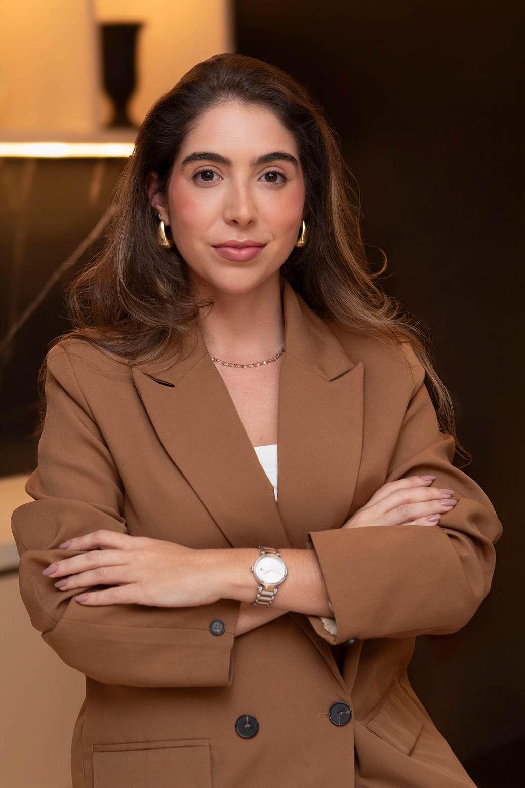 Dra. Débora Guedes de blazer caramelo, braços cruzados, no consultório